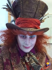 Woman dressed in orange wig, white face makeup and blue eyeshadow wearing a top hat